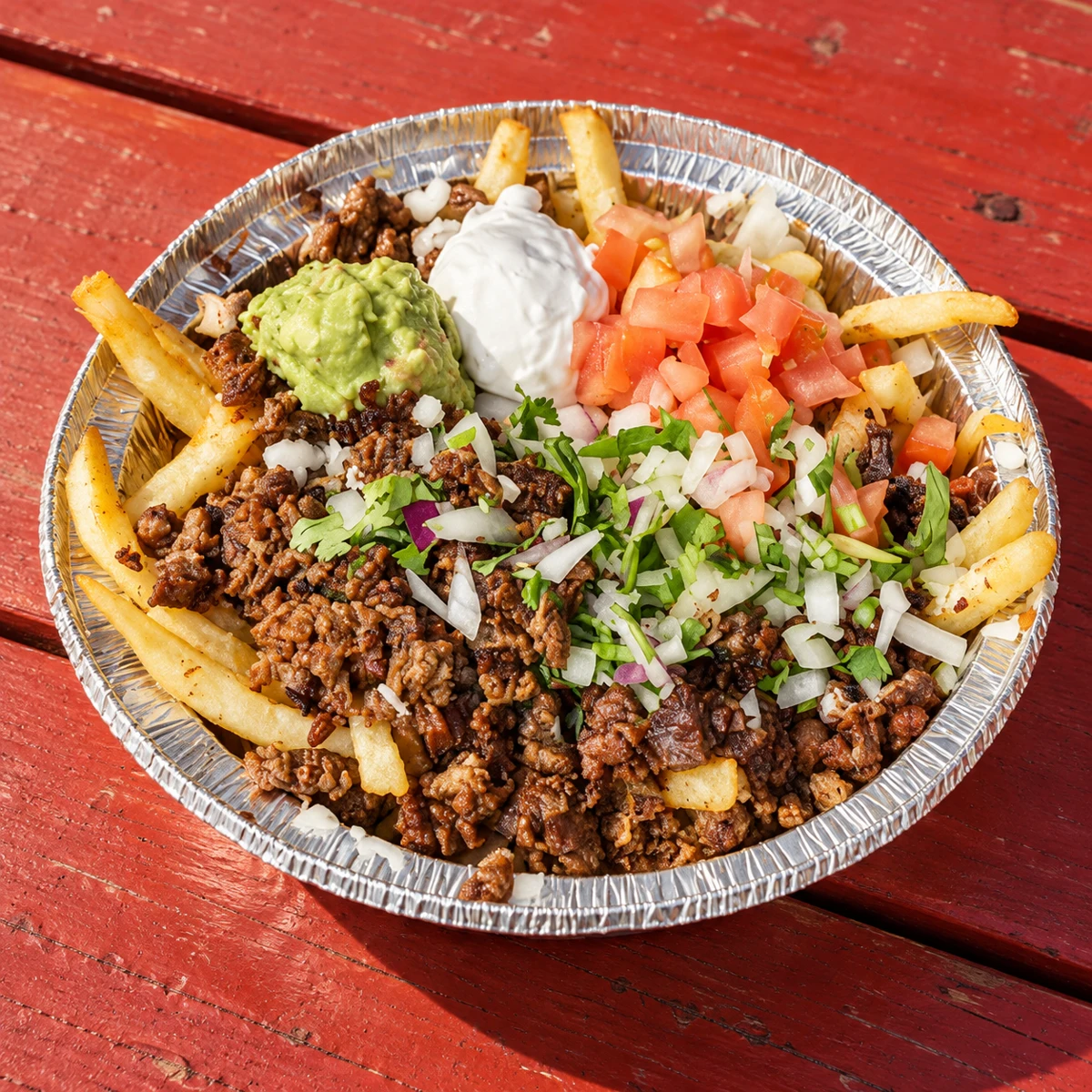 Carne asada fries in a foil tray topped with guacamole, sour cream, tomato, onion, and cilantro